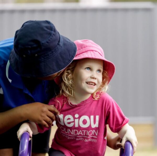 A small child in a pink shirt and hat is shown looking away from the camera, and is being hugged by an educator.