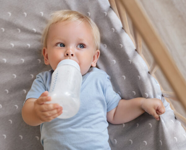A caucasian child is shown lying in a cot, holding a bottle filled with milk.