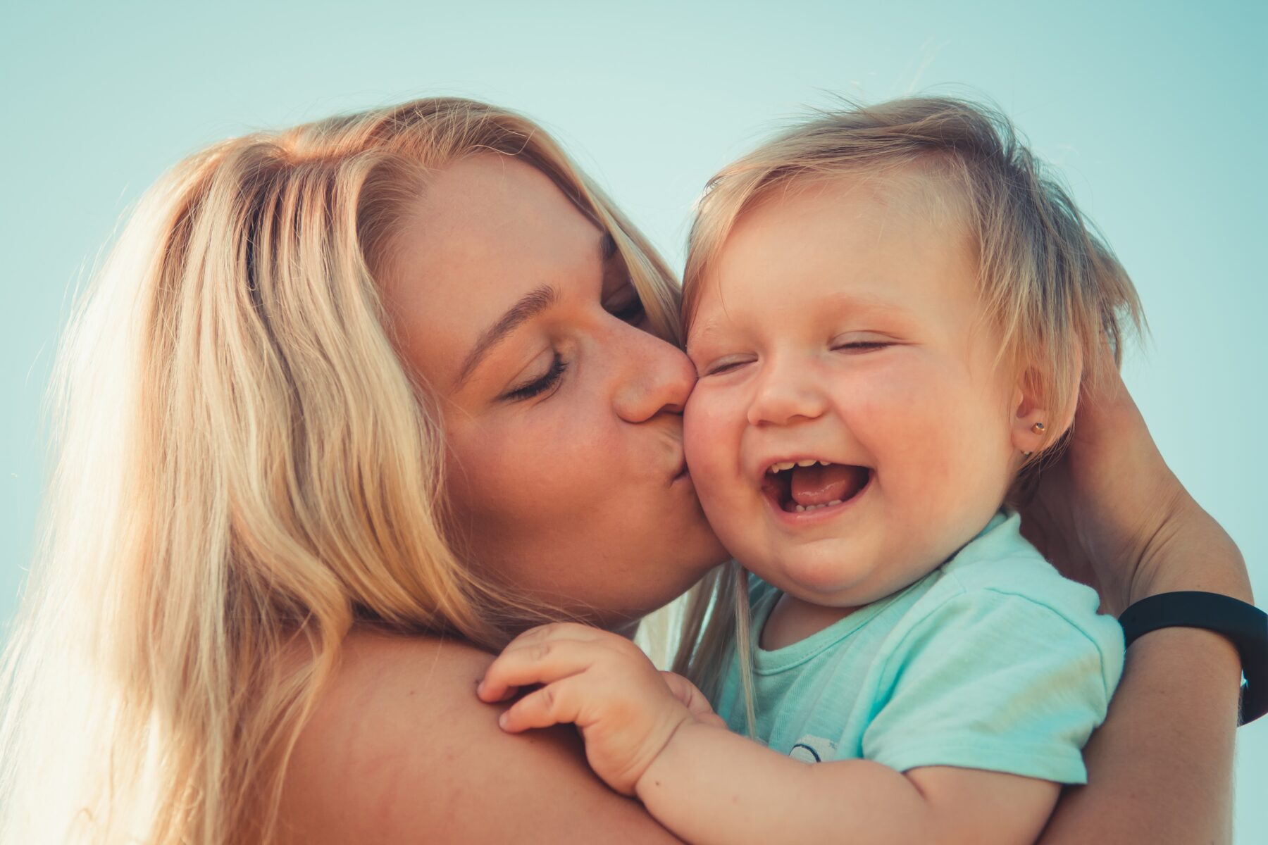 A blonde woman kisses the cheek of a smiling young boy.