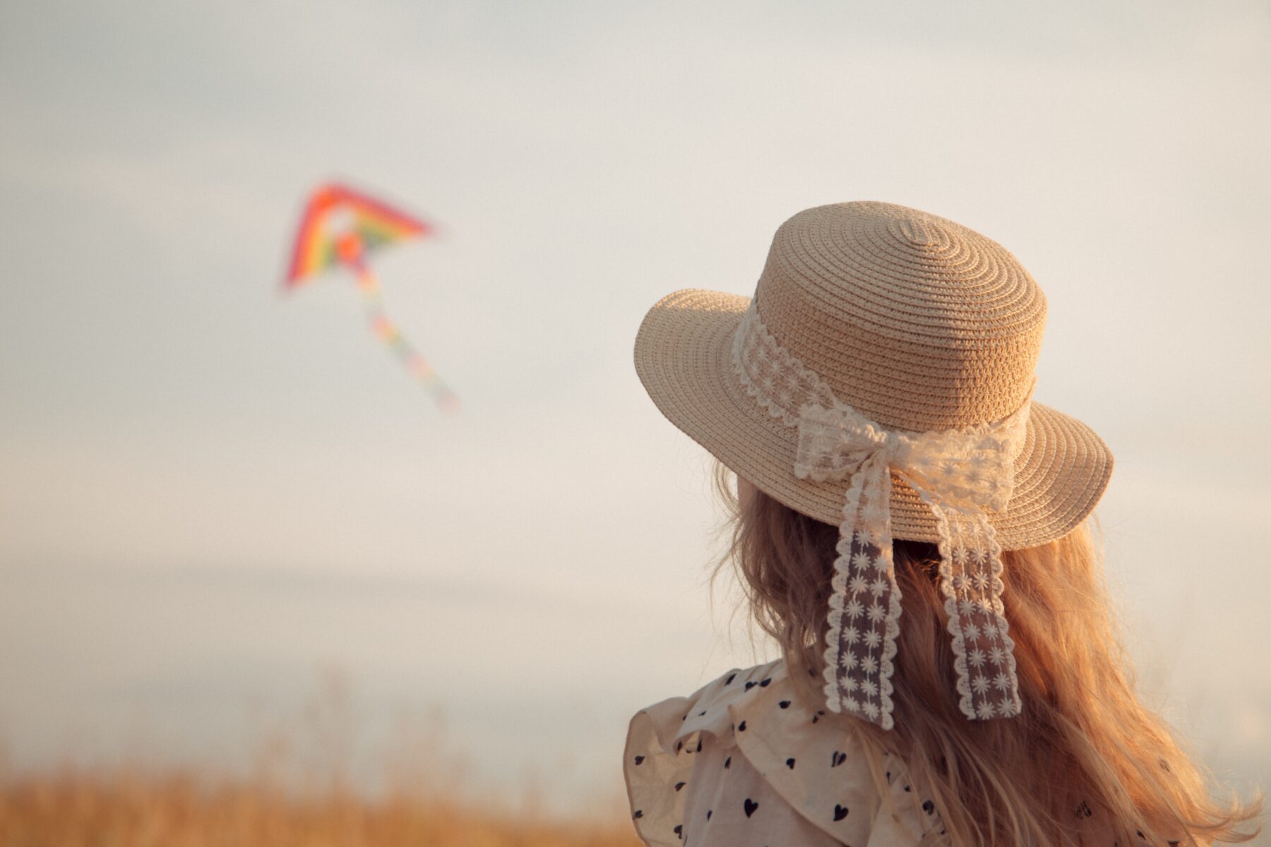 Image shows a girl in a hat with long blonde hair. She faces away from the camera, looking at a kite.